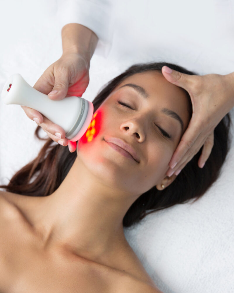 A white woman receiving a red light therapy facial treatment from a technicians hands.