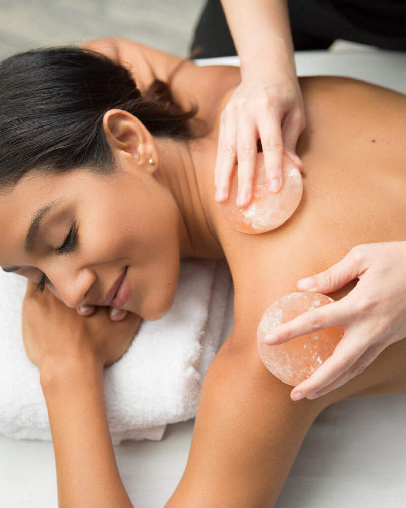 A masseuse's hands placing pink salt stones on a white woman's shoulder.
