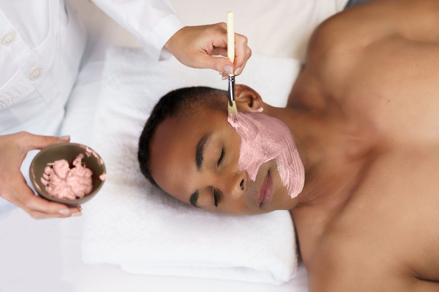A black man lying on a table has a pink facial treatment applied to his face with a brush.